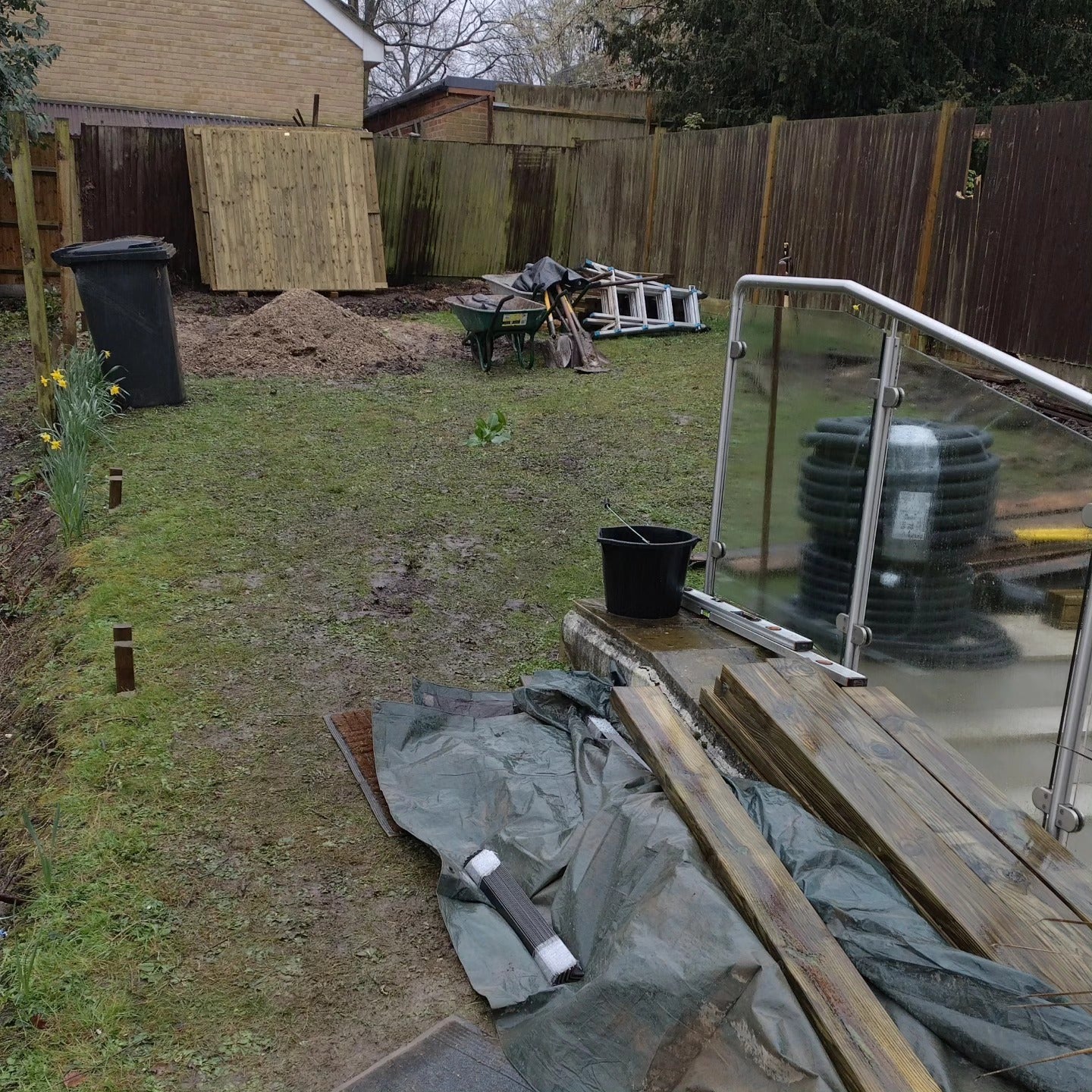 A backyard construction scene with a wooden fence, grassy lawn, a black trash bin, a wheelbarrow, a tarp-covered pile of materials on a wooden deck, and stacked black circular components near a glass railing.